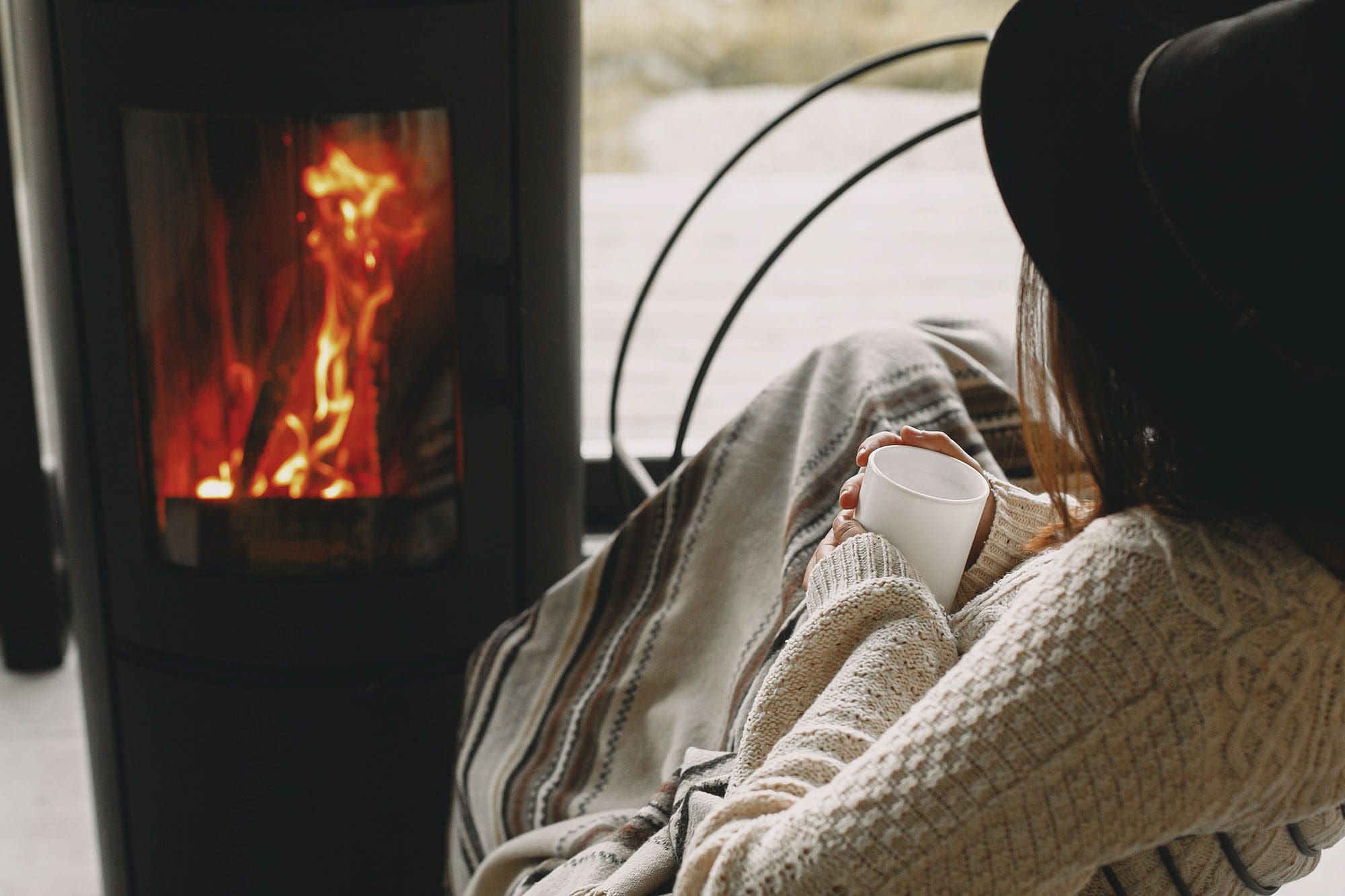 Stylish young woman holding warm cup of tea and relaxing besides a modern black fireplace with view on mountains.