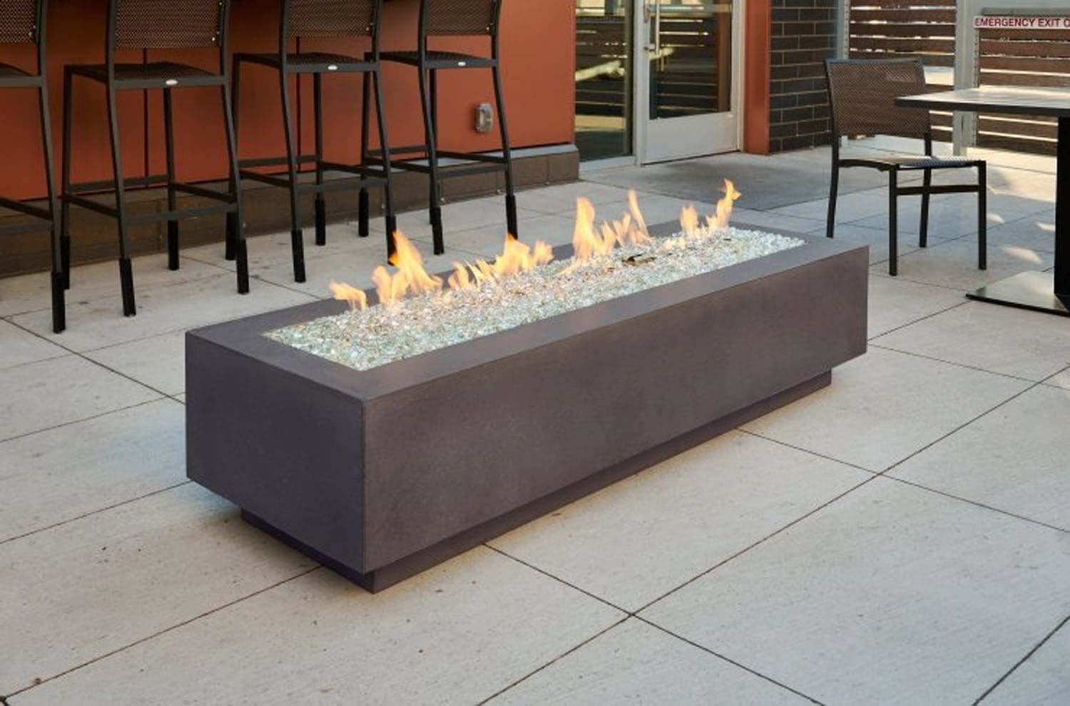 The Outdoor Greatroom Cove 72" Linear Gas Firepit with glass rocks sits on a tiled patio, flames flickering along its length. Bar stools and tables are visible in the background near a building.
