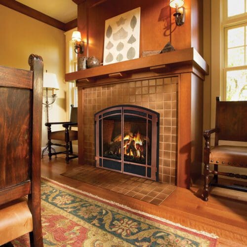 A cozy living room showcases the Mendota DXV gas fireplace series in a tiled surround with a wooden mantel, accent wall art, antique-style wooden chairs, and a patterned rug over hardwood floors illuminated by warm light.