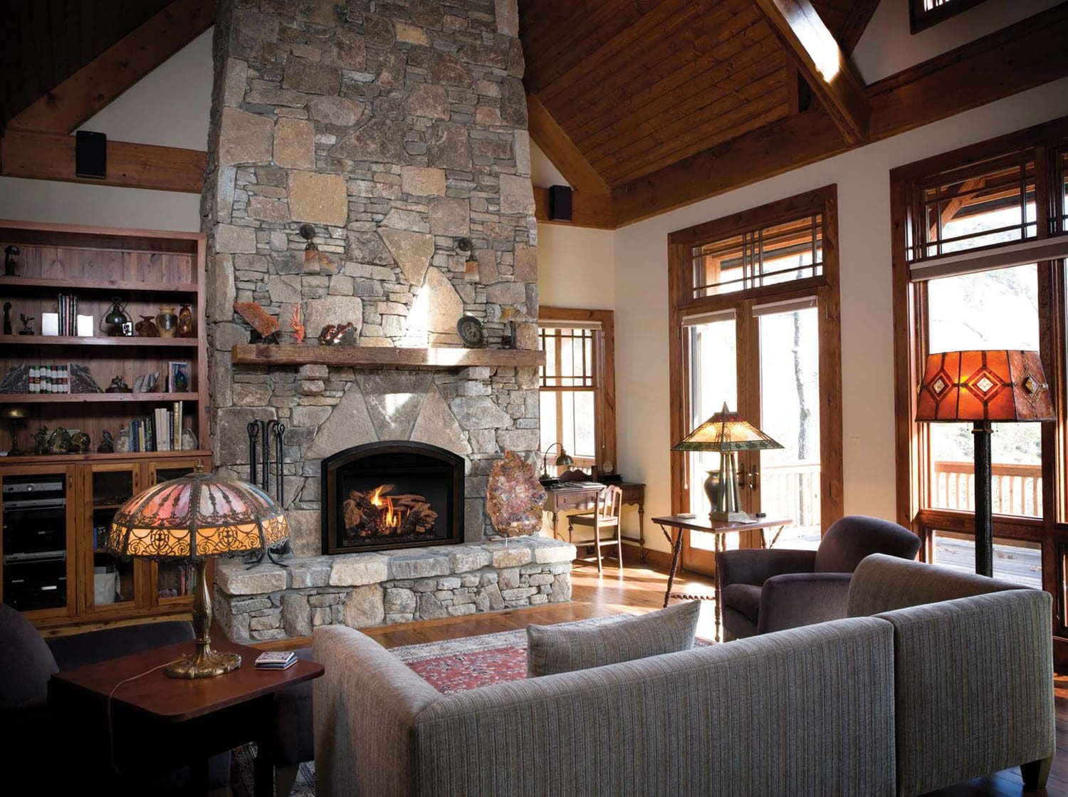 A cozy living room with a large stone fireplace featuring a Mendota FV44i Gas Insert, wood-paneled ceiling, tall windows for natural light, beige sofa, armchairs, stained glass lamps, and built-in wooden shelves filled with decor and books.