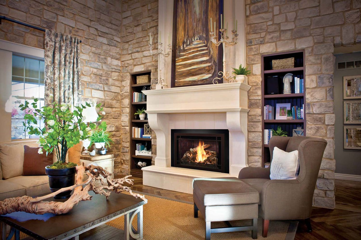 A cozy living room with stone walls features a lit Mendota FV44i Gas insert fireplace, an armchair, sofa, coffee table with a decorative branch, bookshelves, a large painting above the mantle, and a window with curtains letting in natural light.