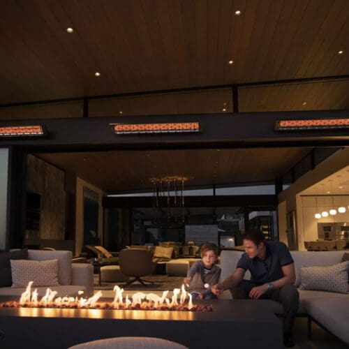 A man and a child sit by a modern outdoor fire pit at night, surrounded by cozy seating and Infratech Comfort Infrared Electric Heaters, with a stylish, warmly lit interior visible in the background.