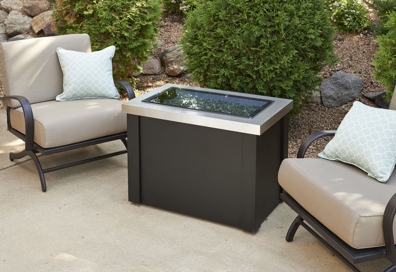 Two beige cushioned patio chairs with patterned pillows flank the Providence Rectangular Gas Firepit Table, arranged on a concrete patio with green shrubs and rocks in the background.