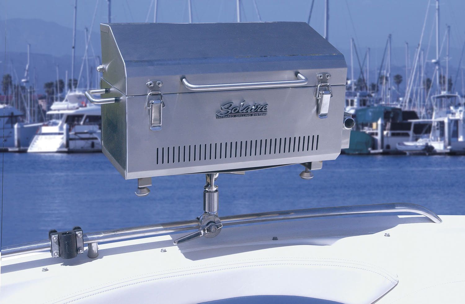 A Solaire Infrared Gas Grill Series stainless steel grill is mounted on a boat railing, overlooking a marina filled with docked sailboats and calm water.