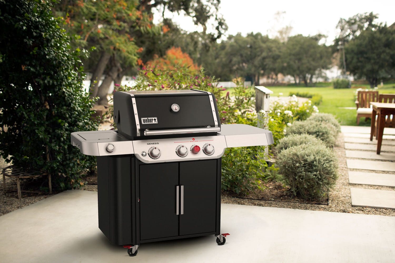 A black and silver Weber Genesis II SP-E-335 gas grill with three control knobs and side tables stands on a patio, surrounded by green shrubs, lawn, and outdoor seating in the background.