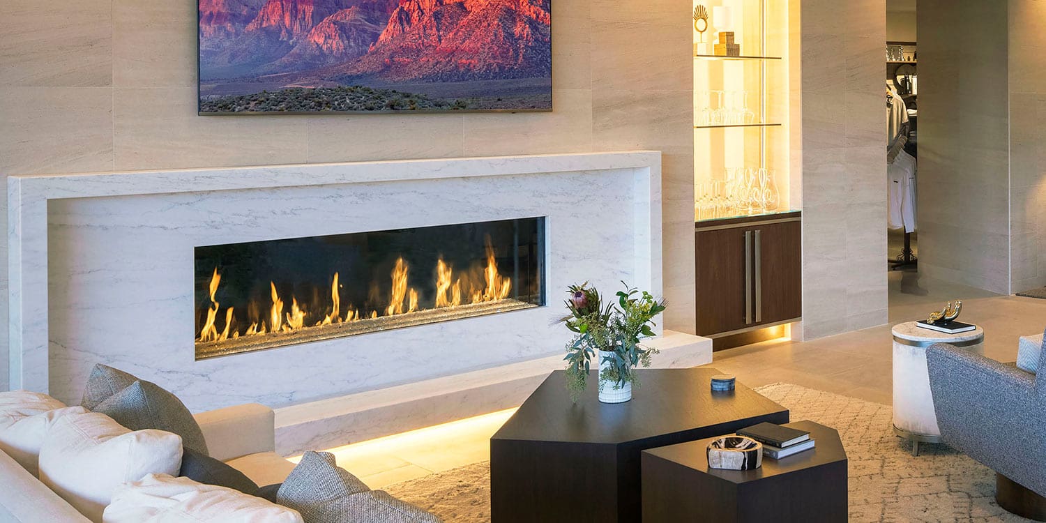 A modern living room features a DaVinci Gas Fireplace set in a white marble wall beneath a TV, with a cozy couch, stylish chairs, a dark coffee table adorned with flowers, and built-in shelves displaying elegant glassware.