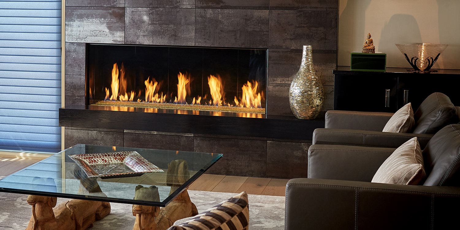 Modern living room with a sleek DaVinci Gas Fireplace, two gray armchairs with striped pillows, a glass coffee table, and decorative accents like a mosaic vase and Buddha statue.