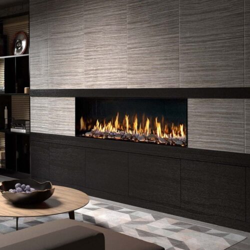 Modern living room with a DaVinci Gas Fireplace in a gray tiled wall, round wooden coffee table, dark sectional sofa, shelves with decor items, and a potted plant to the left.