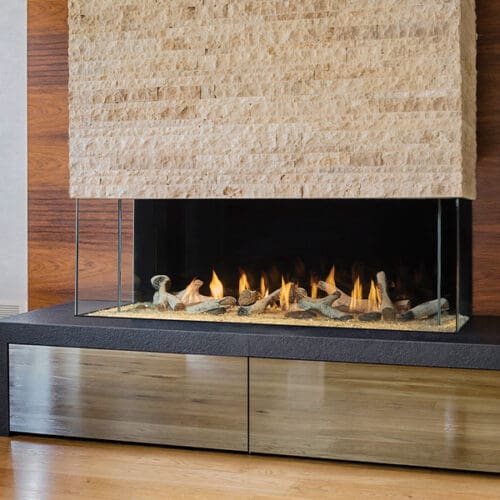 The Firenze™ Collection by DaVinci Custom Fireplaces™ features a single-sided linear gas fireplace with a glass enclosure and decorative stones, built into a wood and stone wall above wooden storage on a polished wood floor.