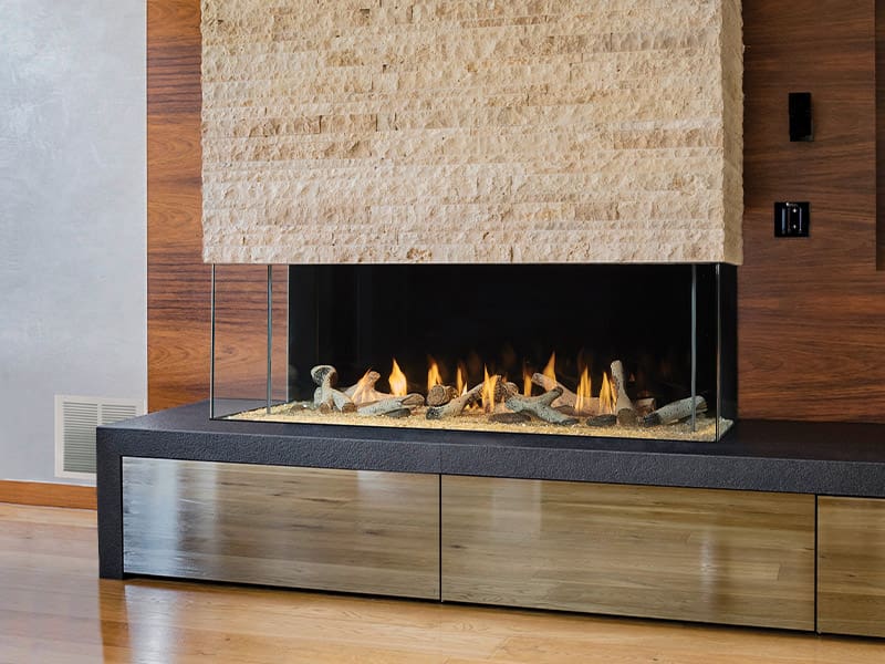 The Firenze™ Collection by DaVinci Custom Fireplaces™ features a single-sided linear gas fireplace with a glass enclosure and decorative stones, built into a wood and stone wall above wooden storage on a polished wood floor.
