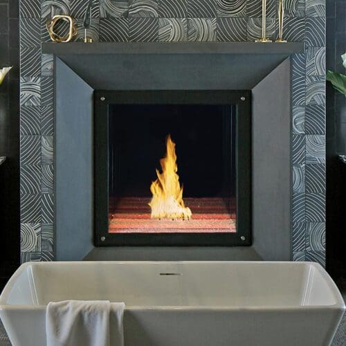 A modern bathroom showcases a freestanding white tub before a Maestro Collection™ By DaVinci Custom Fireplaces, flanked by two vanities with mirrors and flower vases, and a towel draped over the tub.