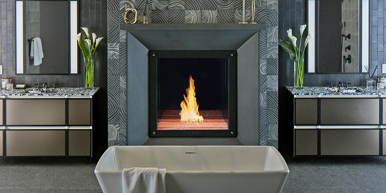 A modern bathroom showcases a freestanding white tub before a Maestro Collection™ By DaVinci Custom Fireplaces, flanked by two vanities with mirrors and flower vases, and a towel draped over the tub.